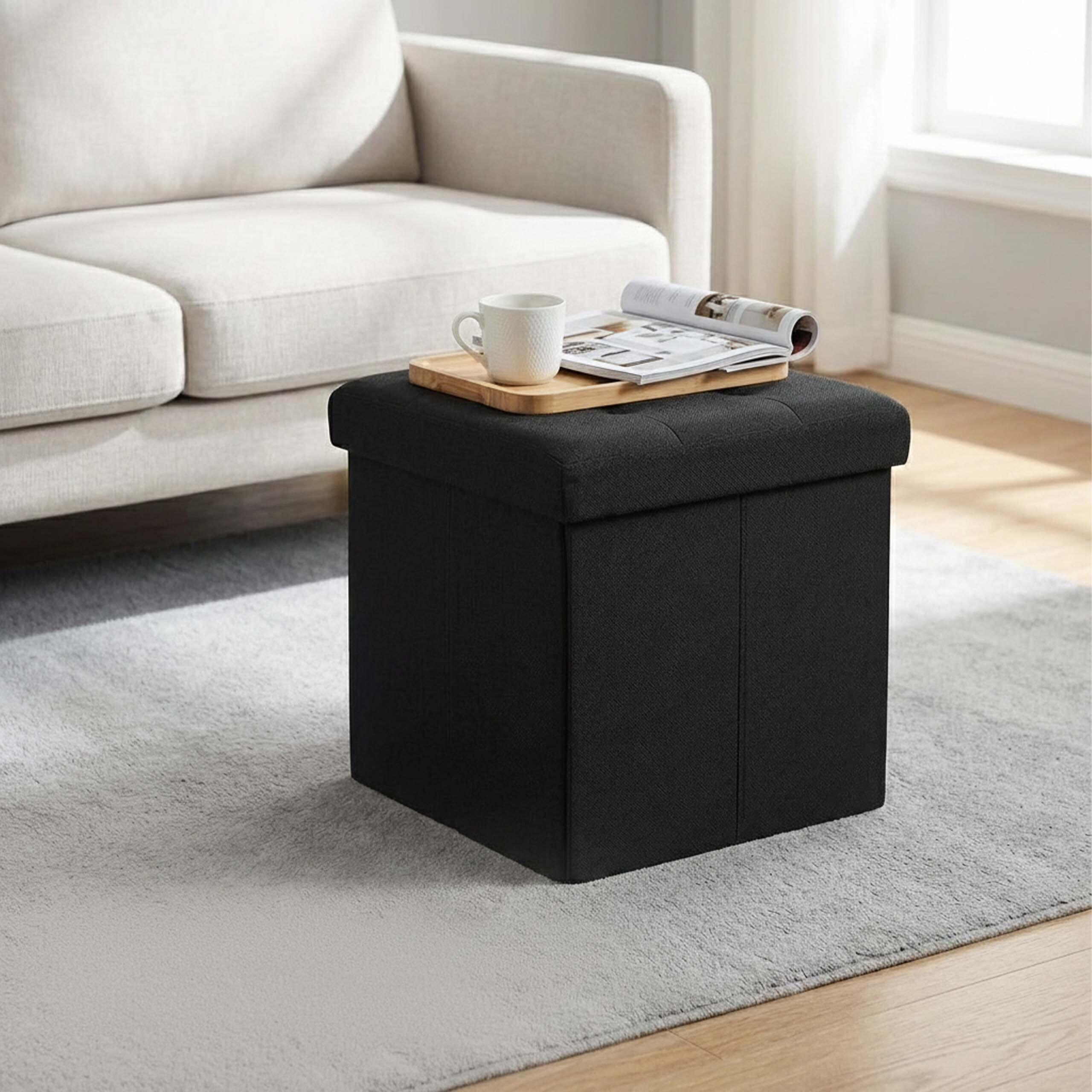 Ein schwarzer quadratischer Hocker mit Holztablett, Tasse und Zeitschrift darauf, auf einem grauen Teppich vor einer Couch.