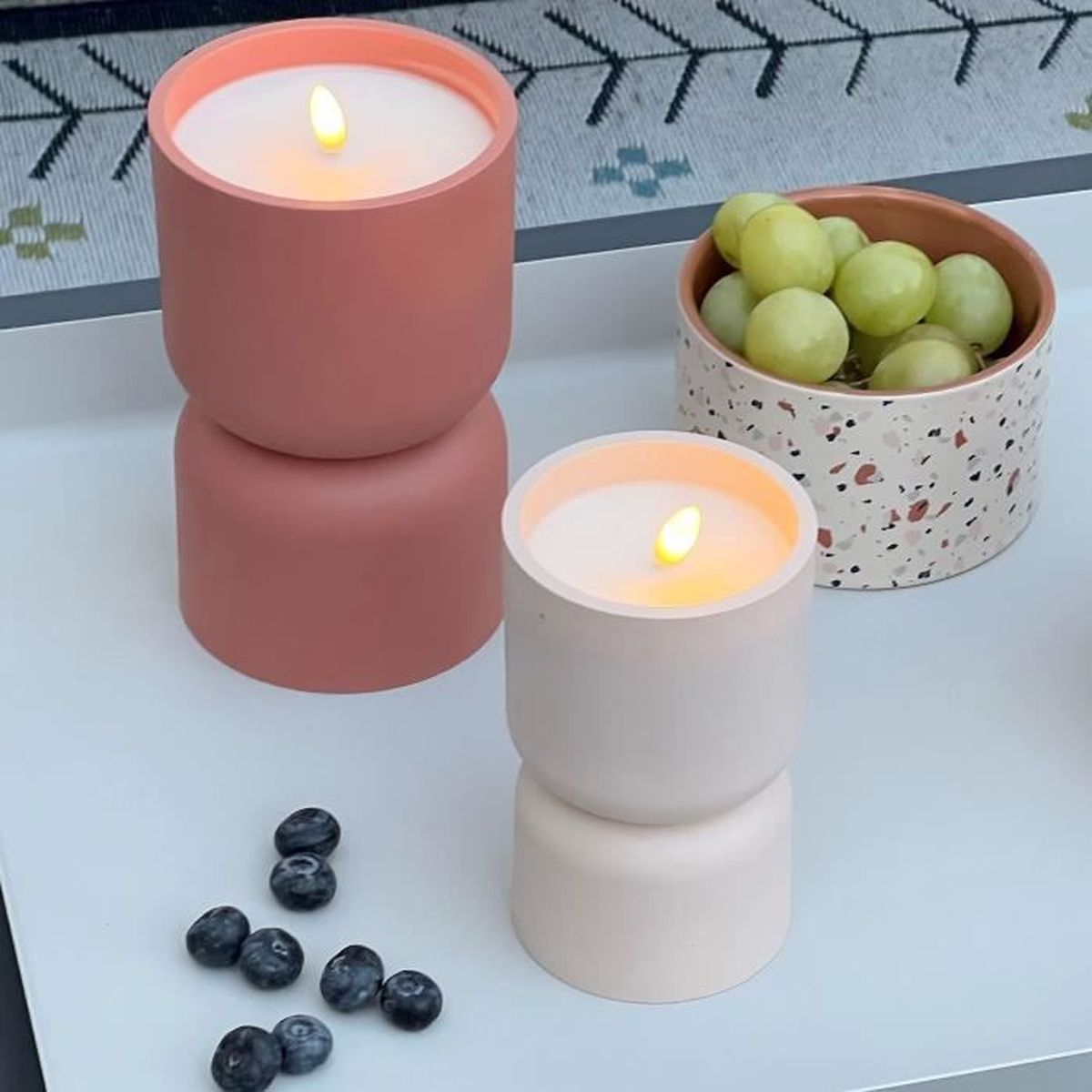 Zwei brennende Kerzen in rosa Haltern, eine Schale mit grünen Trauben und frische Blaubeeren auf einem Tisch.