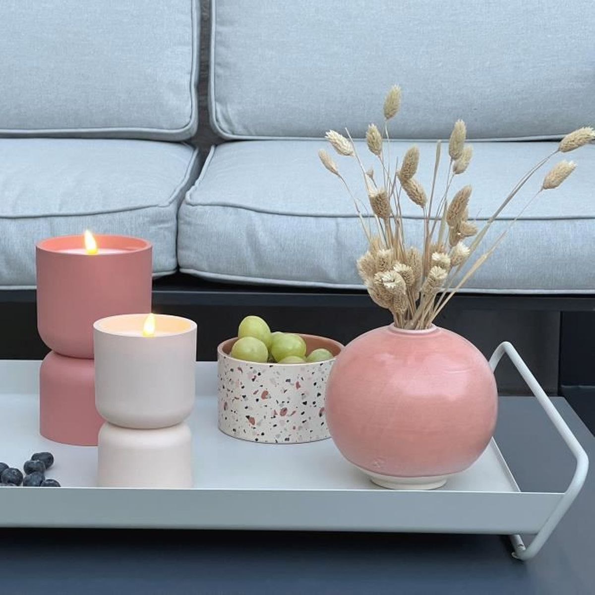 Ein weißes Tablett mit zwei rosa Kerzen, einer Schale voller Weintrauben und einer runden Vase mit Trockenblumen.