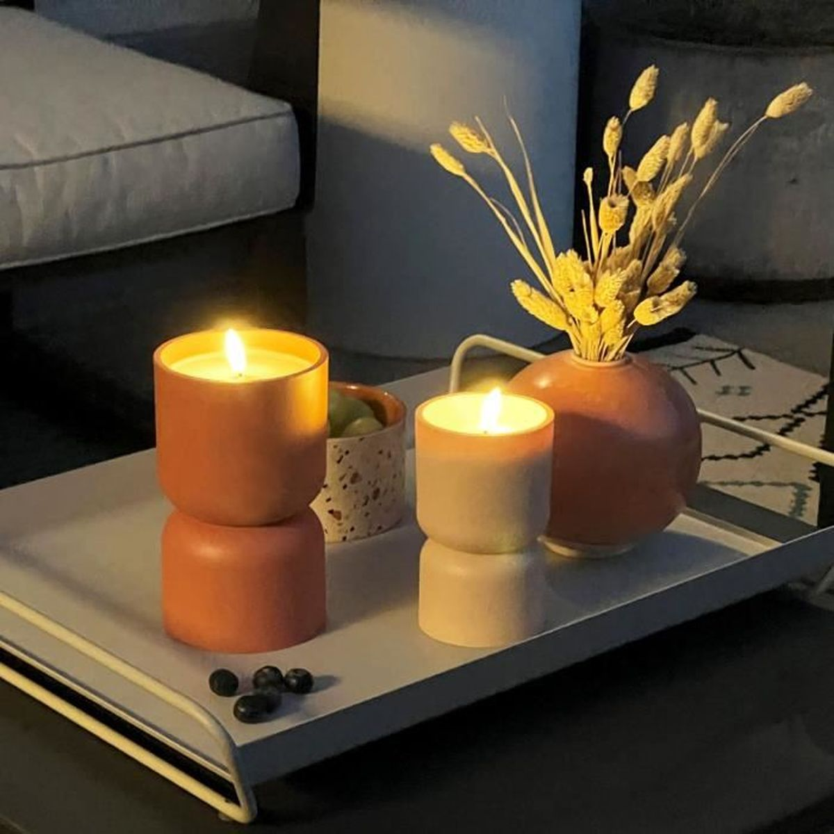 Ein Tablett mit zwei brennenden Kerzen, einer Vase mit Trockenblumen, Beeren und einer Schale auf einem dunklen Tisch.