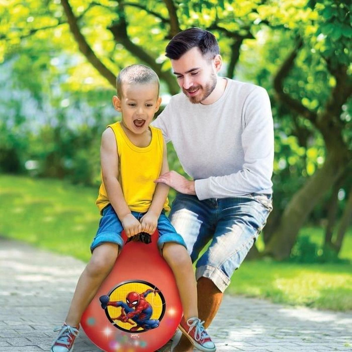 Ein glücklicher Junge hüpft auf einem roten Ball mit Spiderman-Motiv, während sein Vater ihn im Park unterstützt.