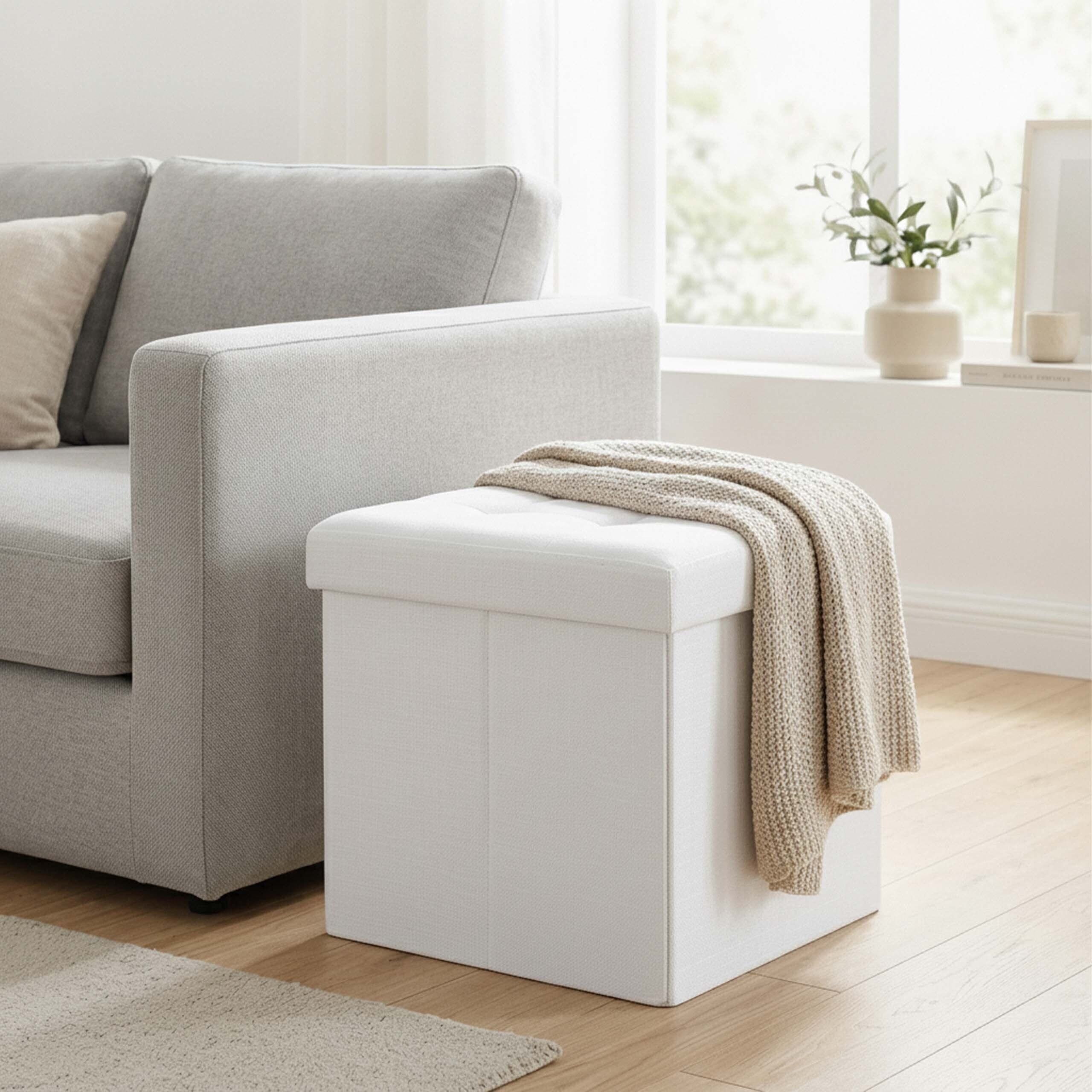Ein weißer Aufbewahrungshocker mit einer beige gestrickten Decke steht neben einem grauen Sofa in einem hellen Wohnzimmer.