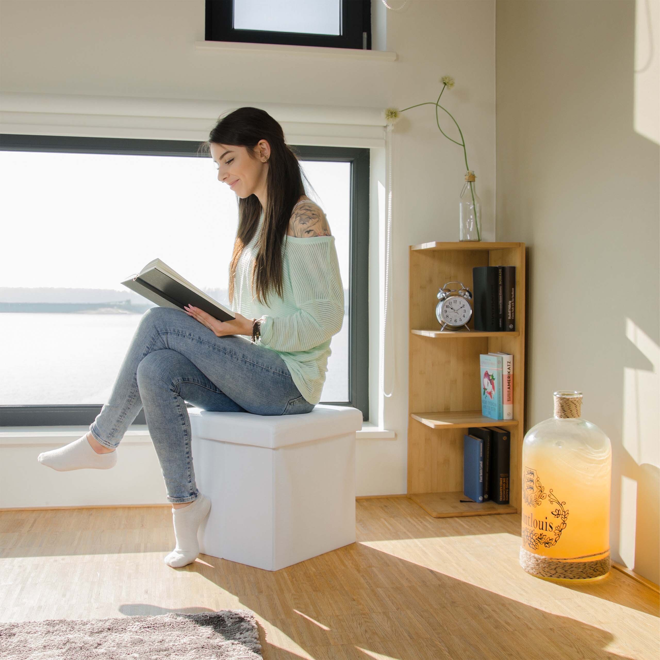 Eine Frau sitzt auf einem weißen Hocker und liest ein Buch am Fenster.