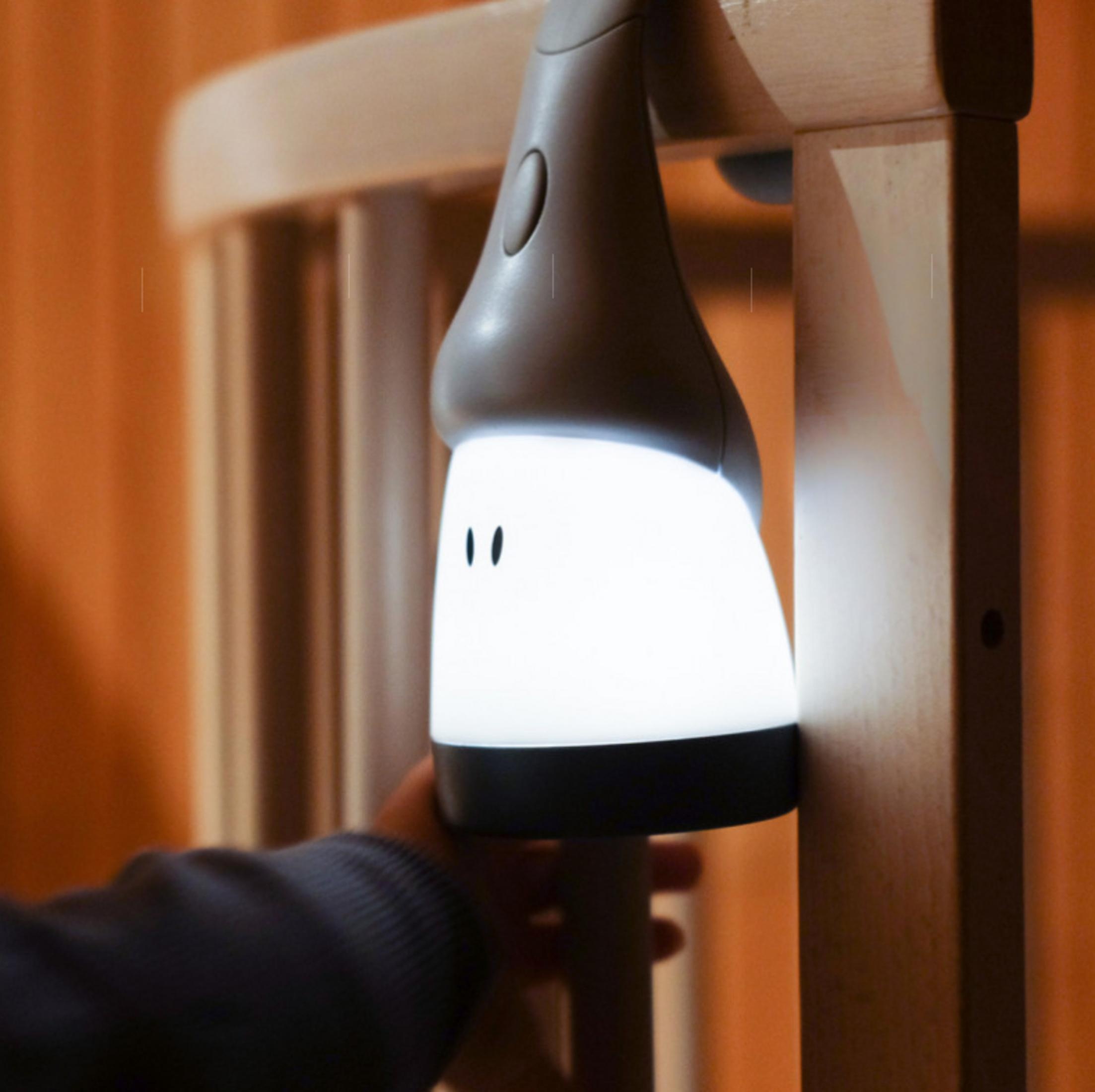 Eine kleine weiße Lampe mit Gesicht hängt an einem Holzbett, eine Hand hält sie.