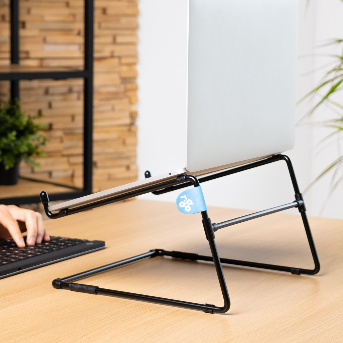 Ein silberner Laptop auf einem schwarzen Metallständer auf einem Holztisch, eine Hand tippt.