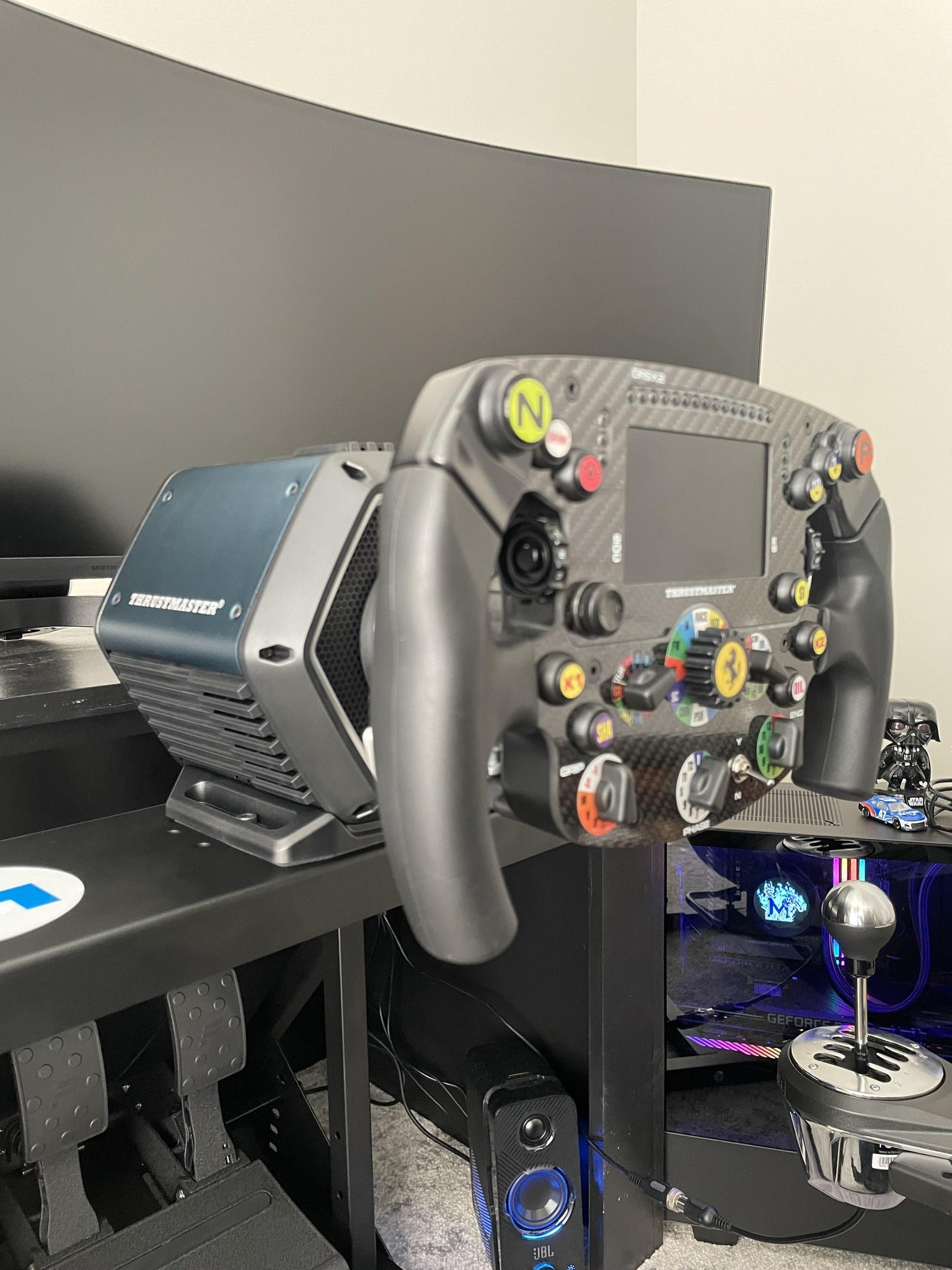 Un volante de carreras Thrustmaster con muchos botones está montado en un escritorio frente a un monitor grande.