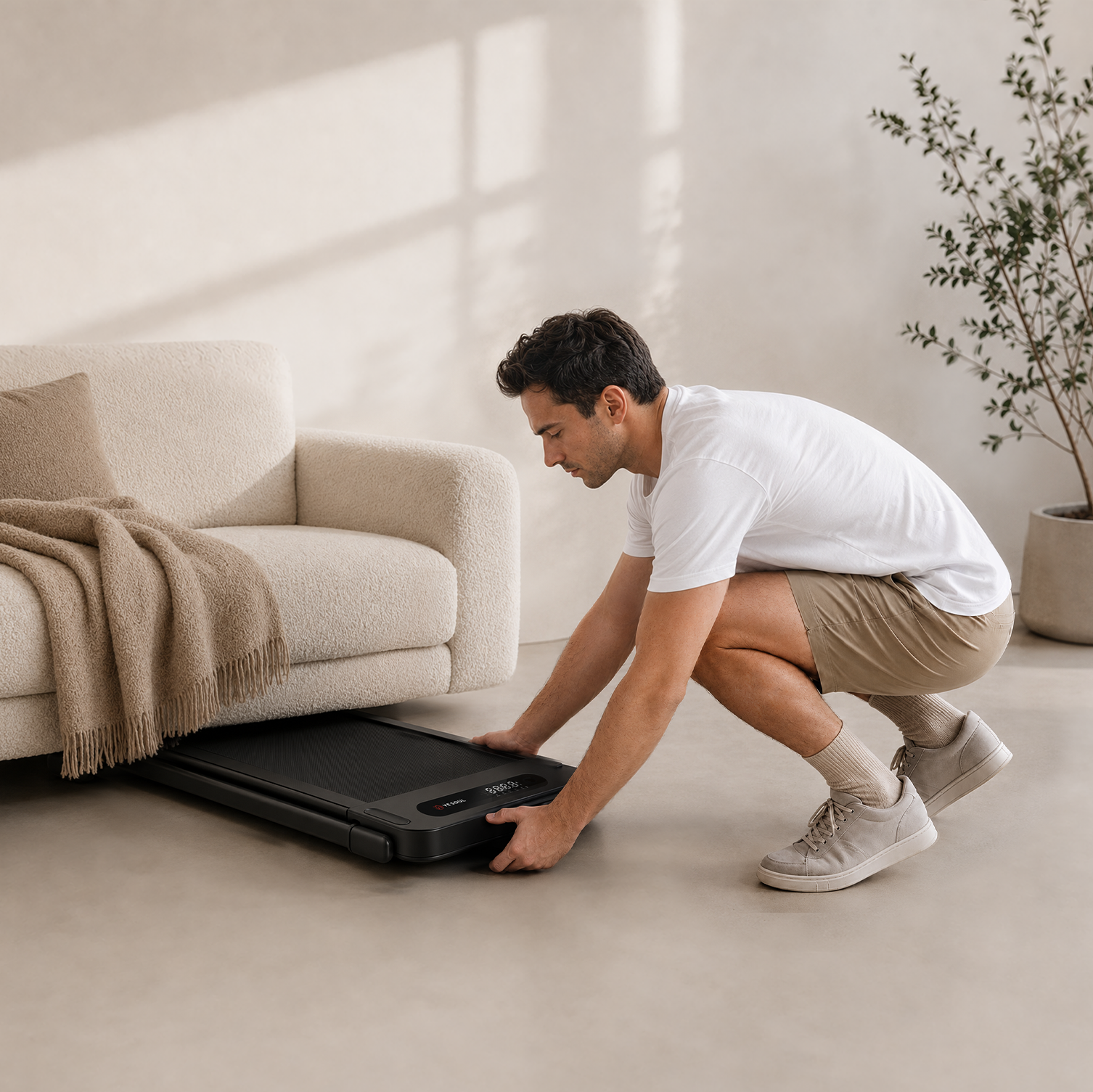 Ein Mann in weißem Hemd schiebt ein schwarzes Laufband unter ein Sofa in einem modernen Zimmer mit Topfpflanze.