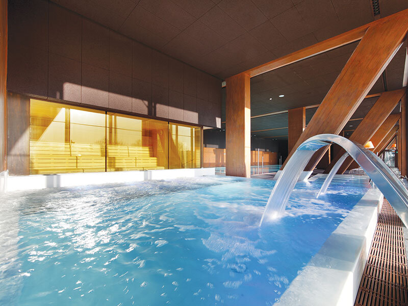 Una piscina cubierta con jacuzzi, juegos de agua y vista a una sauna finlandesa de madera.