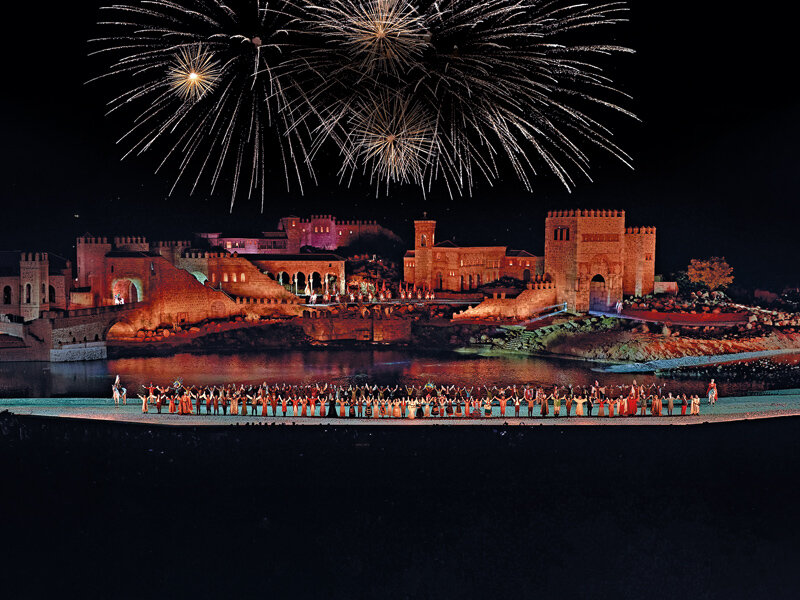 Fuegos artificiales iluminan el cielo nocturno sobre un castillo histórico y una multitud en la orilla.