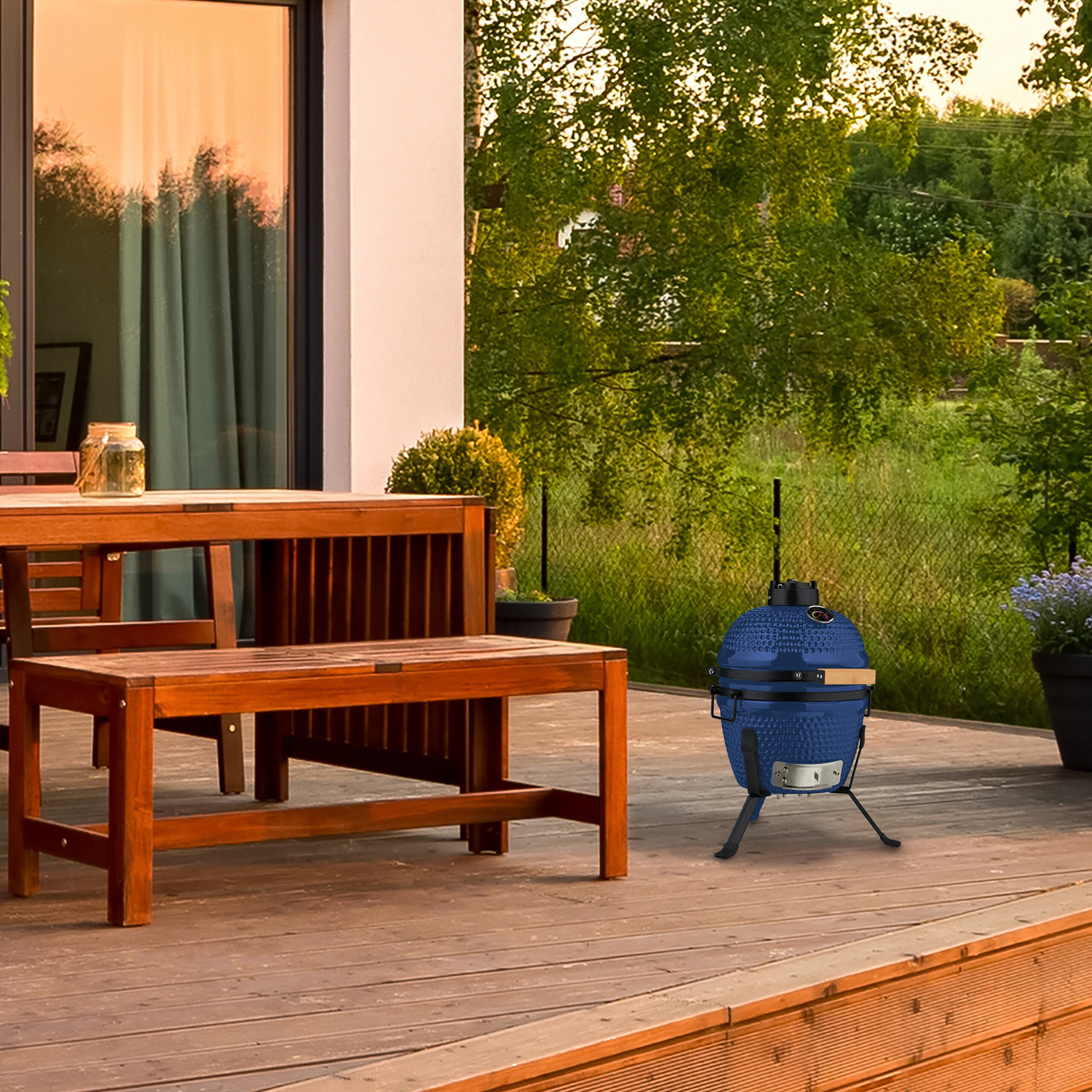 Ein blauer Keramikgrill steht auf einer Holzterrasse neben einem Tisch und Bänken, im Hintergrund Bäume.