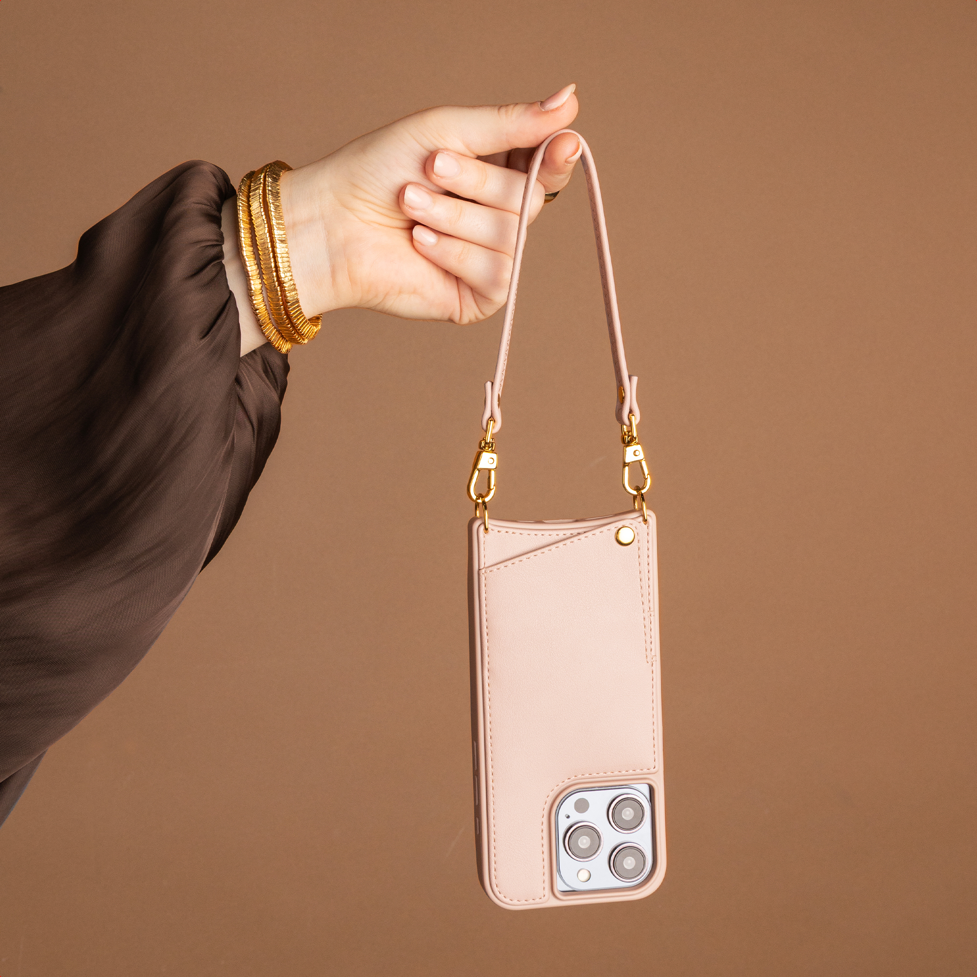Hand hält ein pastellrosa Handycase mit langem Band und Goldakzenten vor braunem Hintergrund.
