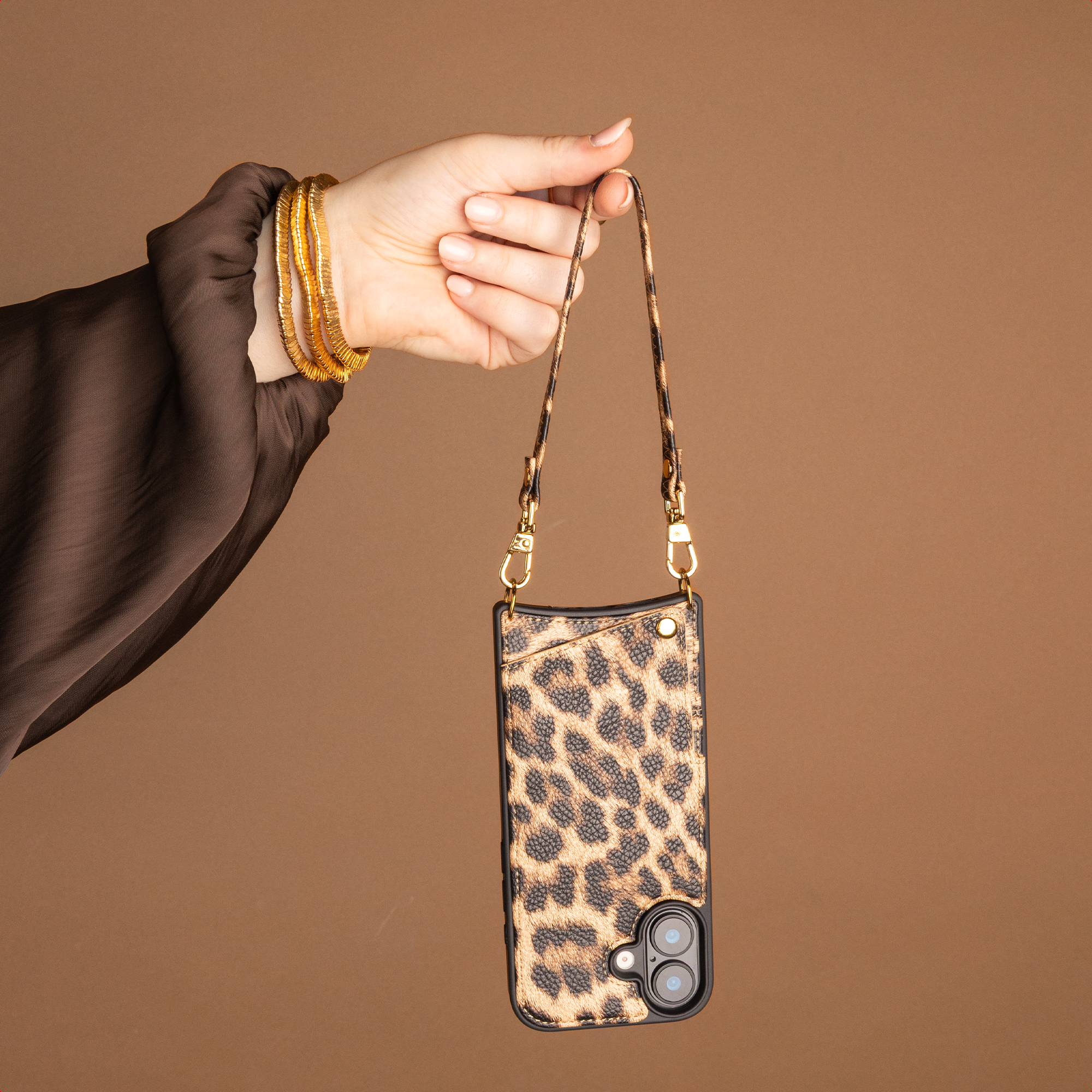 Eine Hand mit goldenen Armreifen hält eine Handyhülle mit Leopardenmuster an einem passenden Riemen vor braunem Hintergrund.