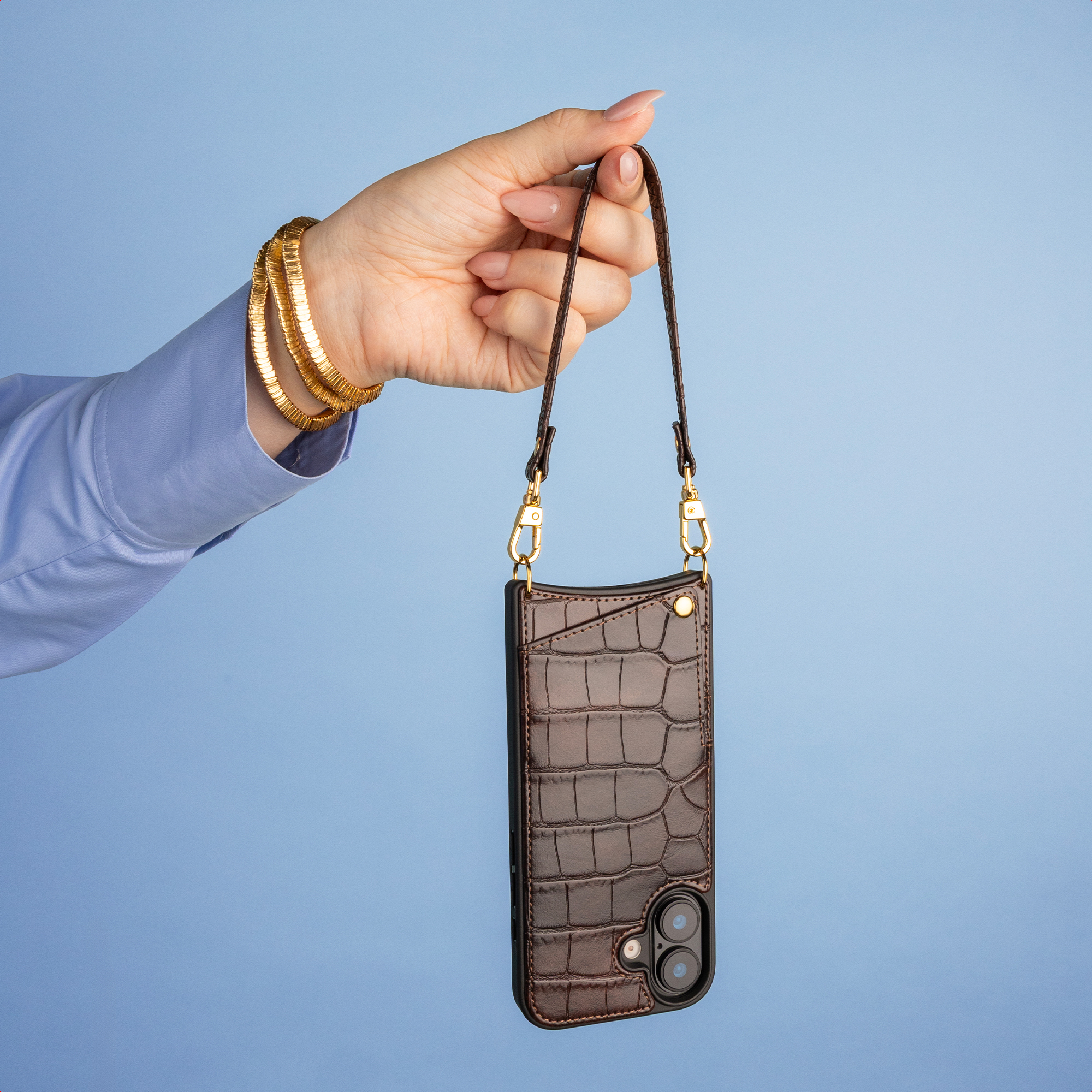Hand holds a brown crocodile-pattern leather phone case with a strap against a blue background.