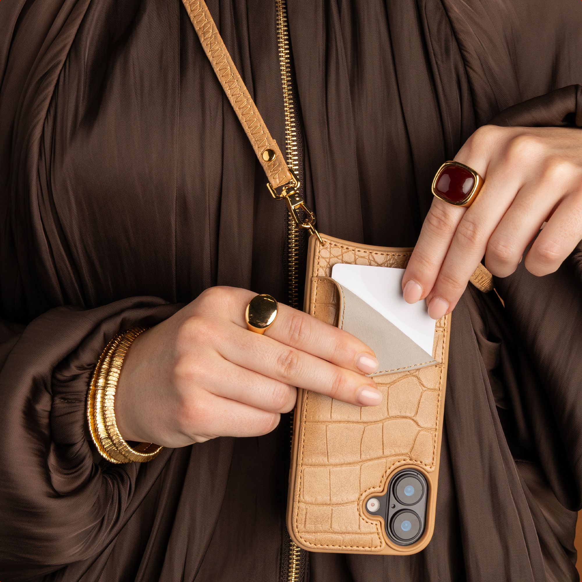 A person in a brown dress inserts a white card into a tan, croco-patterned phone case with a matching strap.