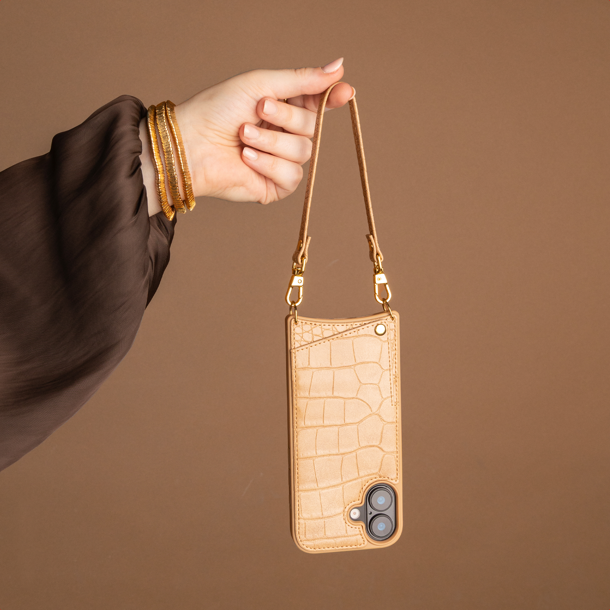 A hand wearing gold bangles holds the strap of a beige, crocodile-textured phone case against a brown background.