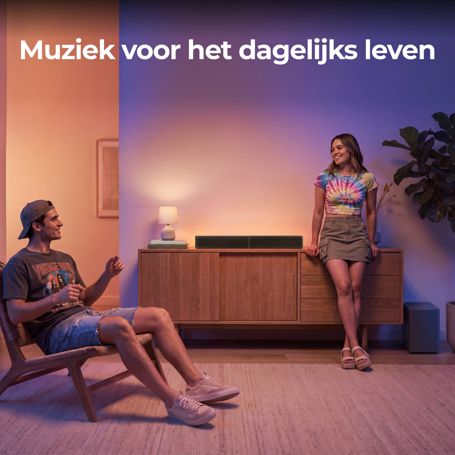 Een jonge man zit in een stoel en lacht naar een vrouw leunend op een houten dressoir met een soundbar.