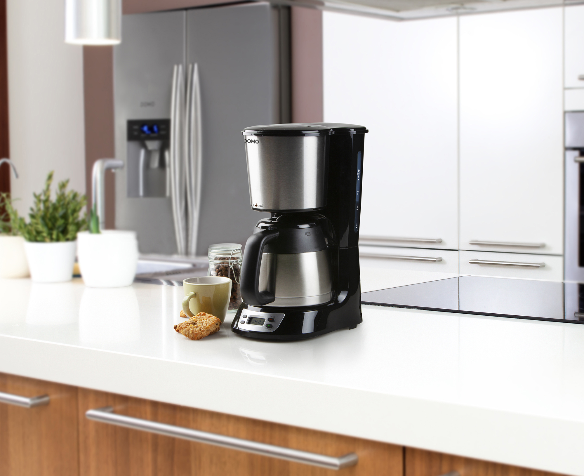 Une cafetière avec verseuse isotherme est posée sur un plan de travail blanc dans une cuisine moderne.