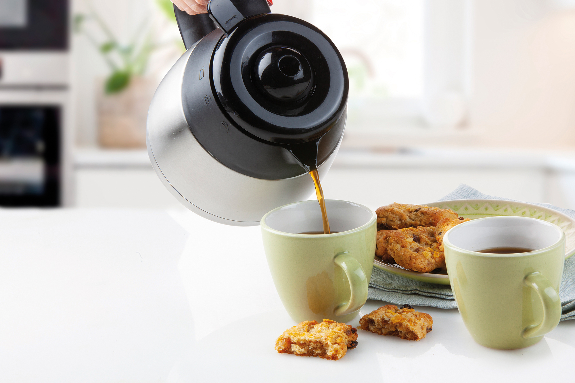 Du café est versé d'une cafetière argent ée dans une tasse verte, avec des biscuits.