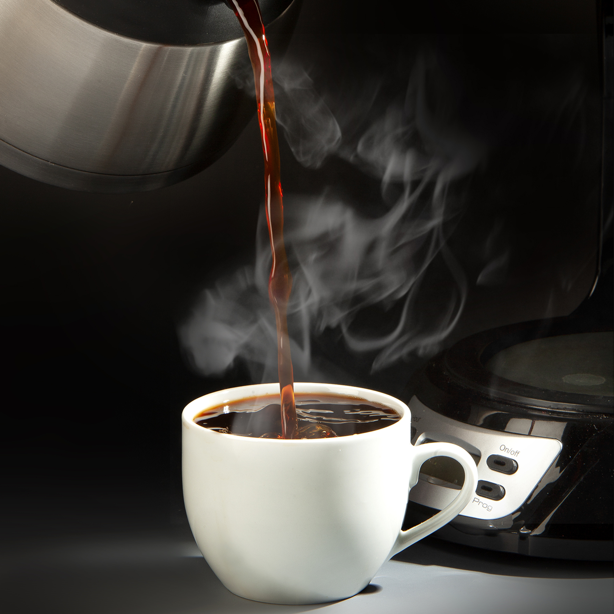Du café chaud est versé d'une cafetière dans une tasse blanche, avec de la vapeur sur fond sombre.