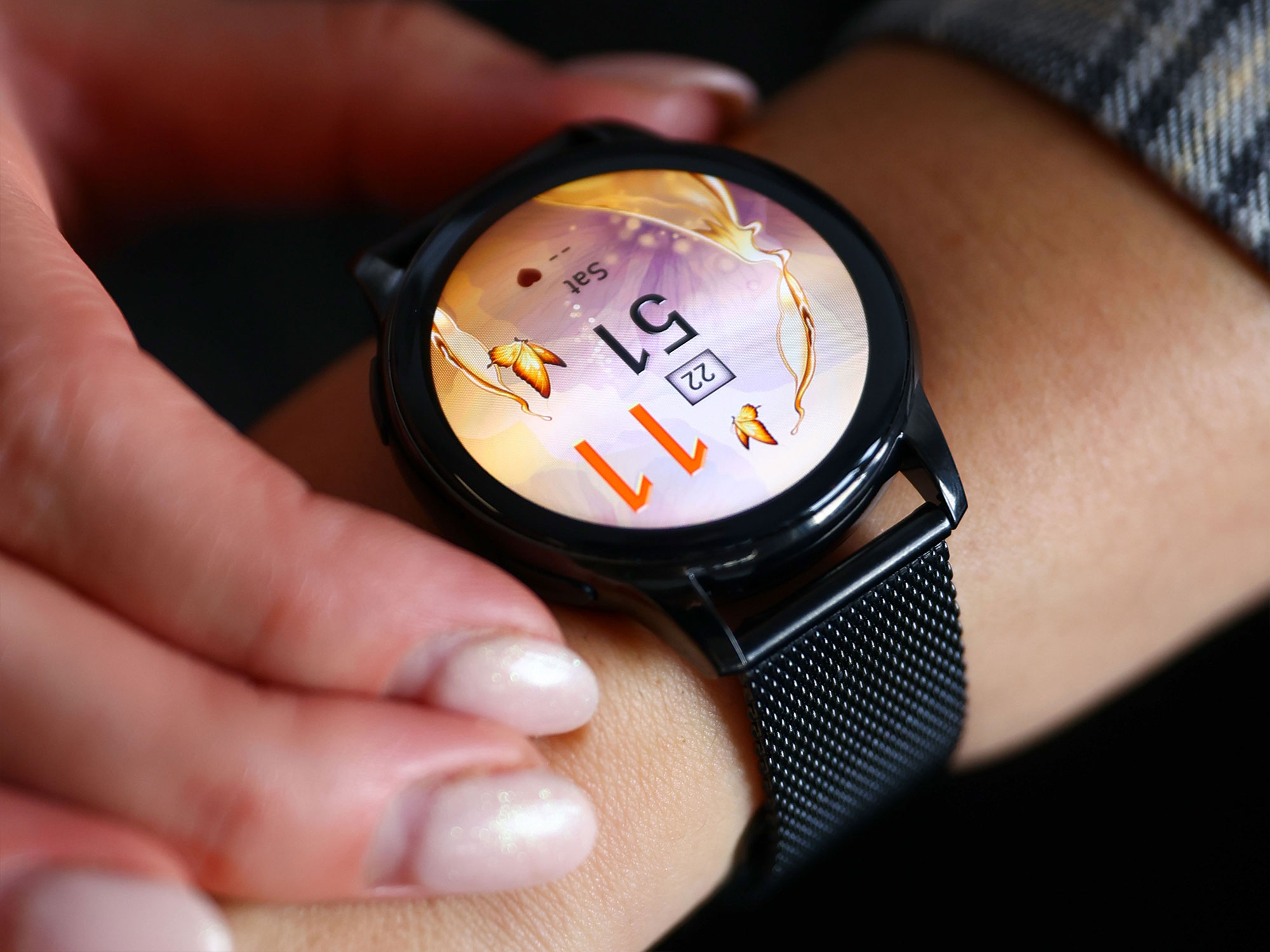 Dłoń nosi czarny smartwatch z kolorową tarczą i metalową bransoletą siatkową.