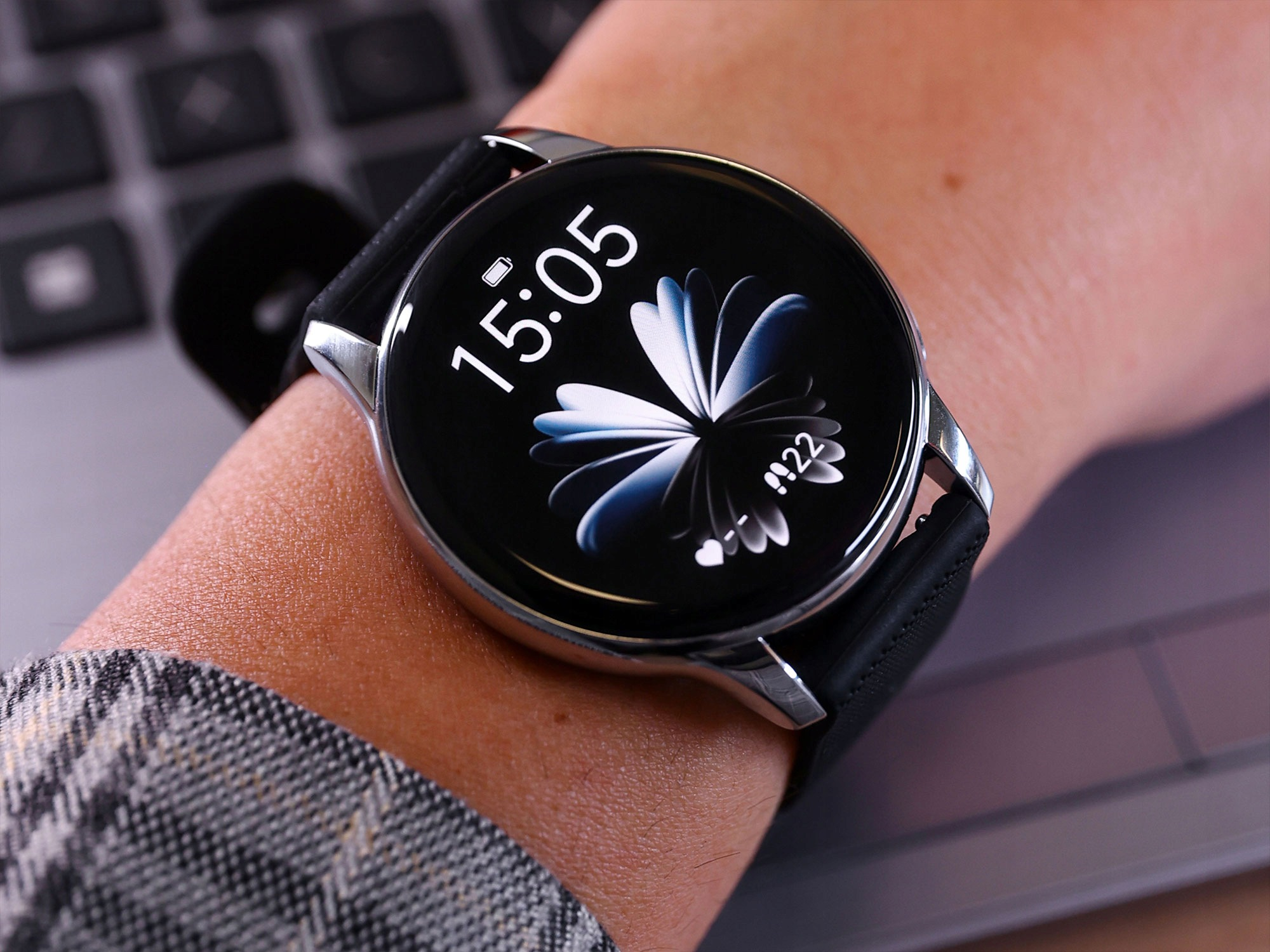 Okrągły smartwatch z czarnym wyświetlaczem pokazuje godzinę 15:05, założony na nadgarstek na tle klawiatury laptopa.