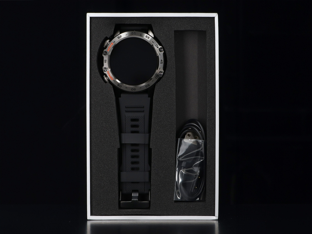 Czarny smartwatch z metalową ramką i paskiem w pudełku obok kabla USB.