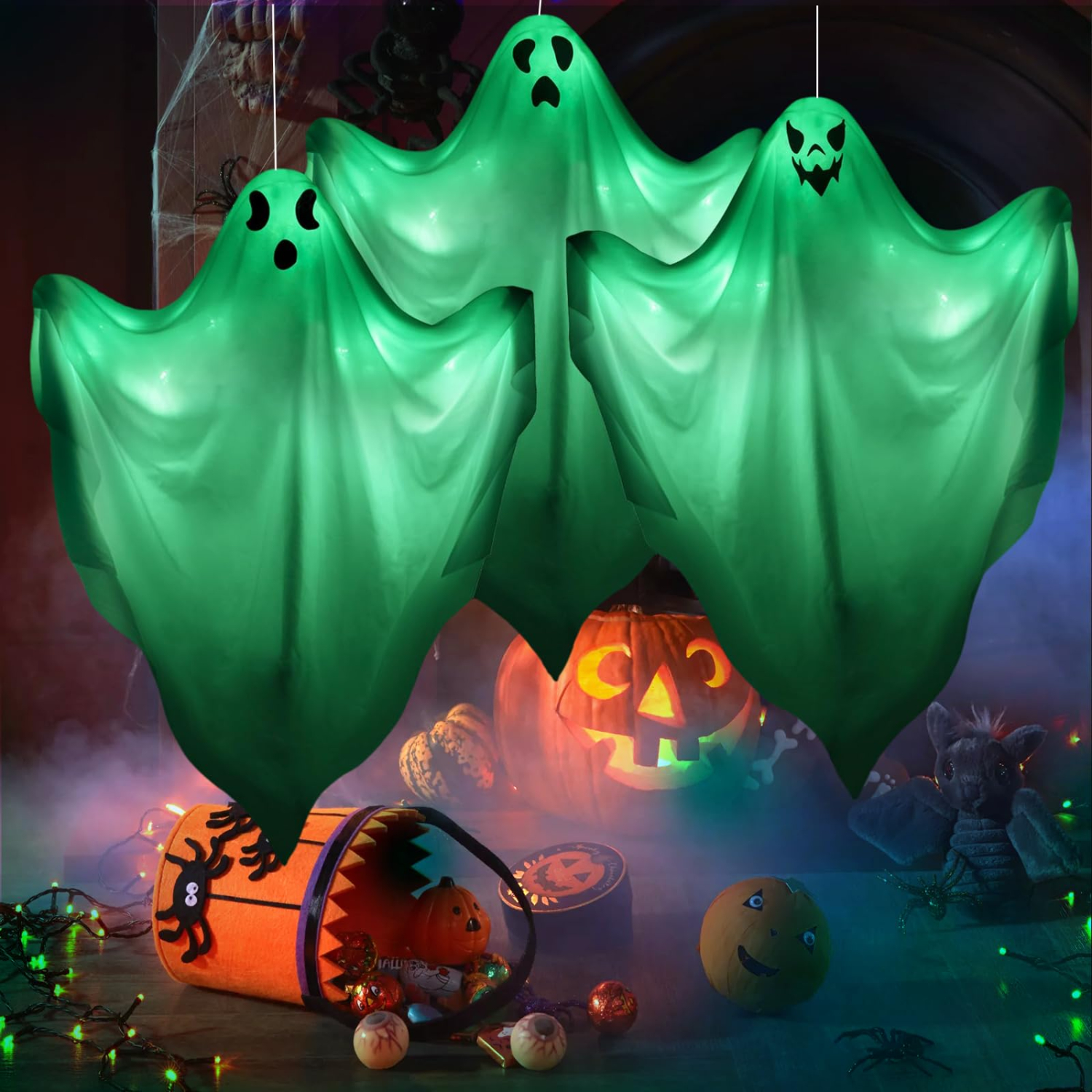 Drei grüne, leuchtende Hängedekorations-Geister schweben über Halloween-Kürbissen und einer Süßigkeitentasche.