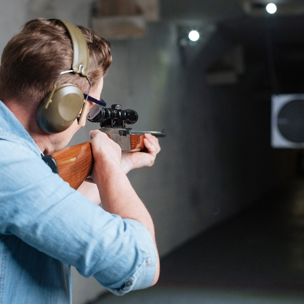 Ein Mann mit Gehörschutz und Brille zielt in einer Schießstandhalle mit einem Gewehr auf eine Zielscheibe.