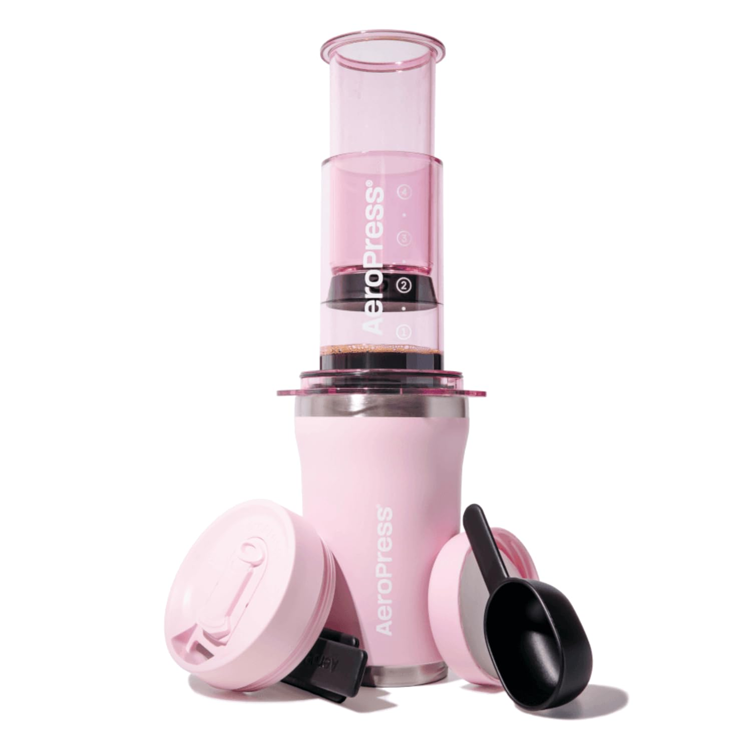 Różowy ekspres do kawy AeroPress na kubku z pokrywką i czarną łyżeczką, na białym tle.