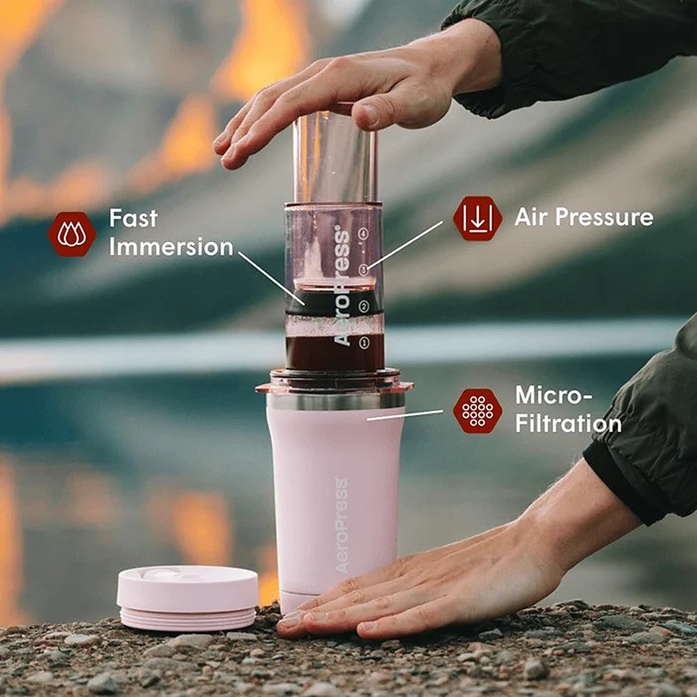Osoba używa ekspresu AeroPress na skalistym podłożu, z etykietami tekstowymi.