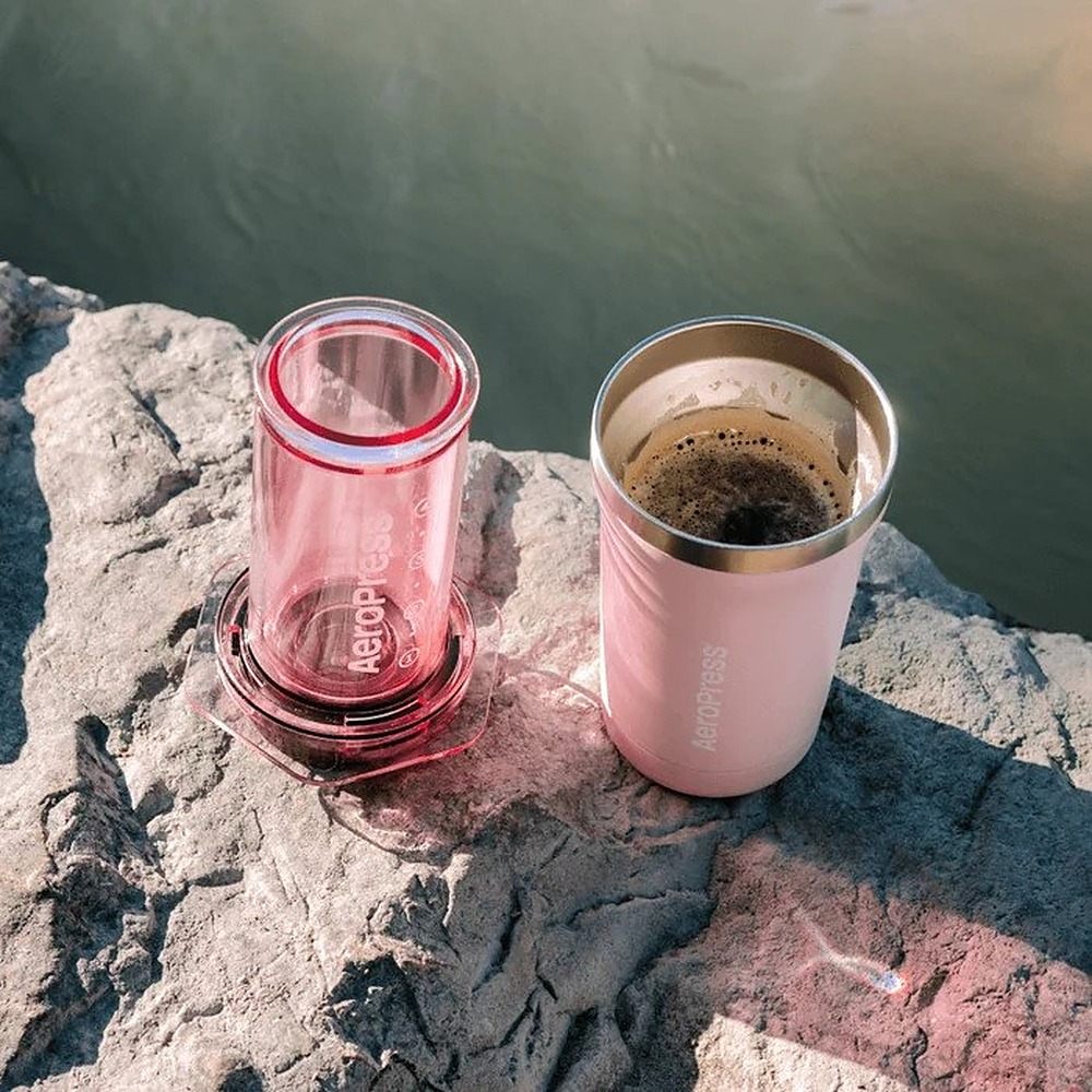 Różowy AeroPress i pasujący kubek kawy stoją na skale nad wodą.