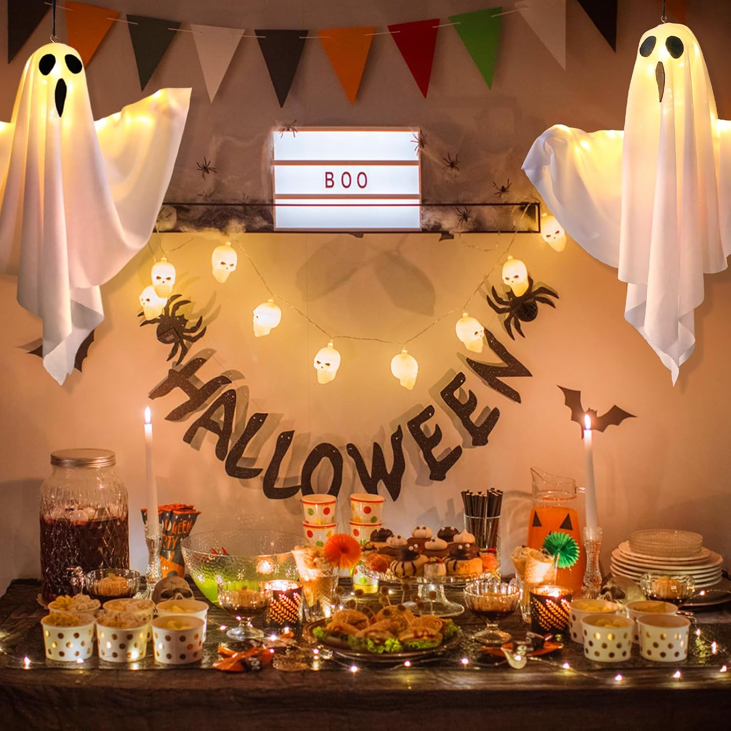 Ein festlicher Halloween-Partytisch mit leuchtenden Geist-Dekorationen, Totenkopf-Lichterkette, Leckereien und BOO-Schild.