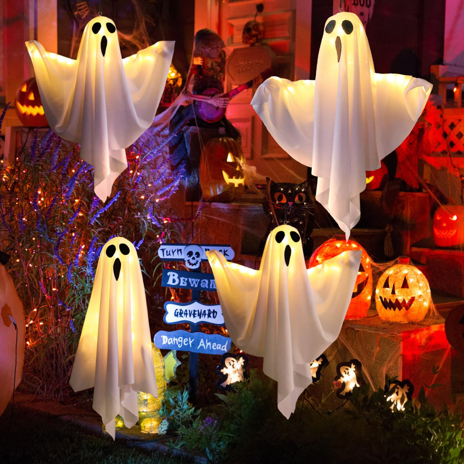 Vier beleuchtete weiße Geisterdekorationen in einem Garten vor einem Haus mit Kürbissen und Schildern zu Halloween.