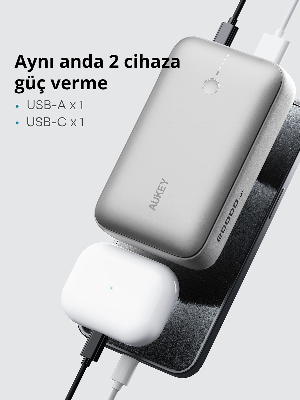 Gri bir Aukey taşınabilir şarj cihazı ve beyaz kulaklıklar USB kablolarıyla aynı anda şarj ediliyor.