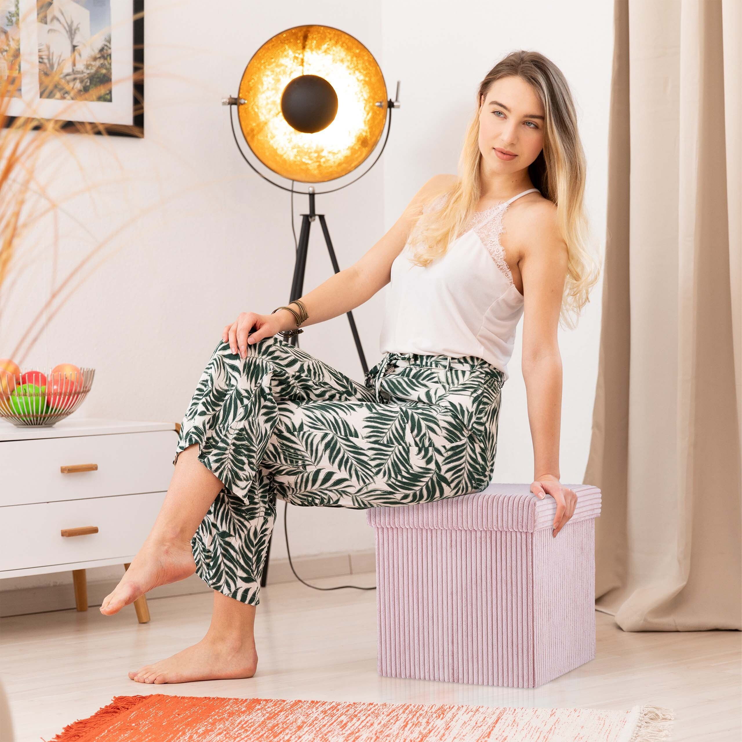 Eine blonde Frau in gemusterten Hosen sitzt auf einem rosa Kocker in einem hellen Raum mit Lampe.