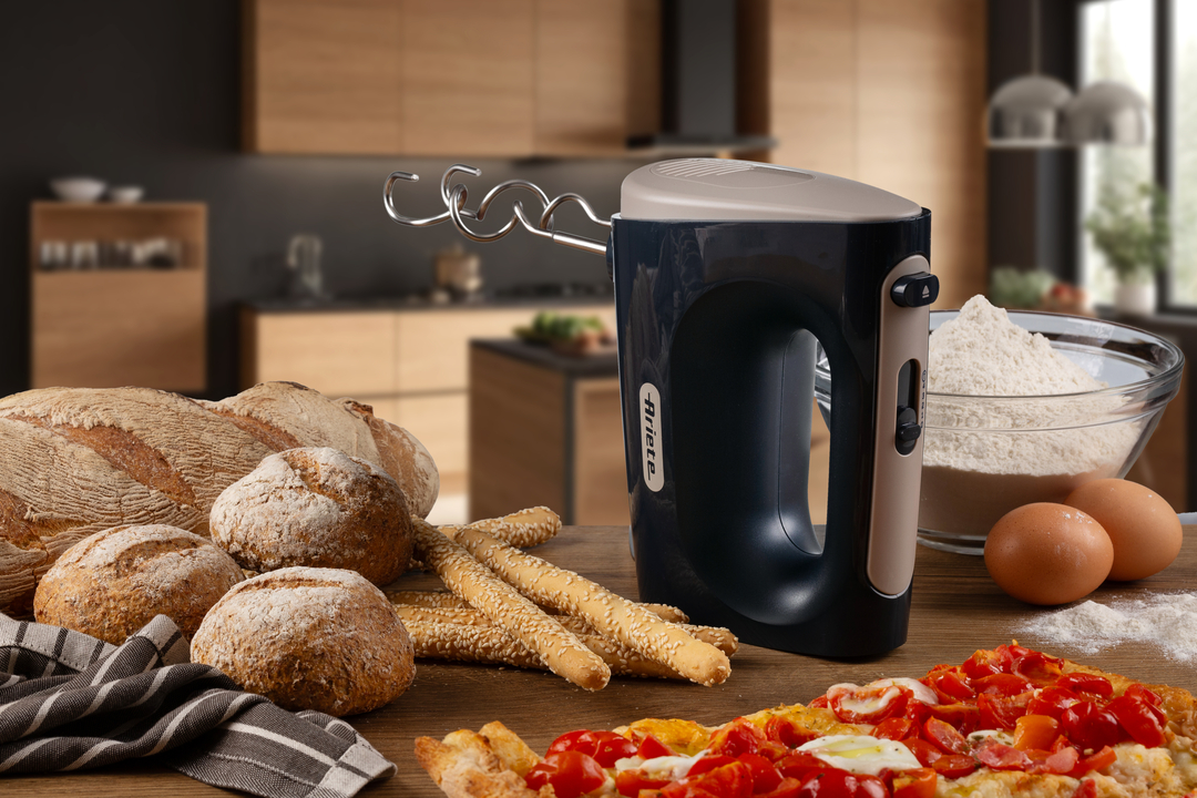 Hand mixer on a wooden table with bread, flour, eggs, and pizza in a kitchen.