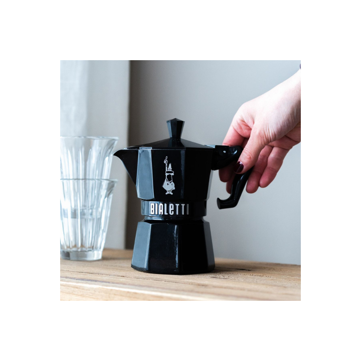 Una mano tiene una moka Bialetti nera su un asse di legno accanto a dei bicchieri.