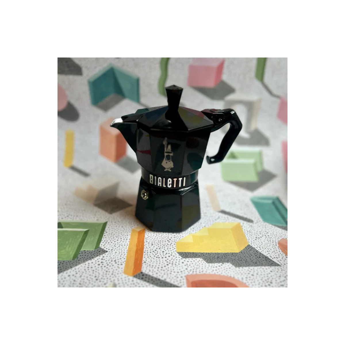Una moka Bialetti nera su sfondo con forme geometriche colorate.