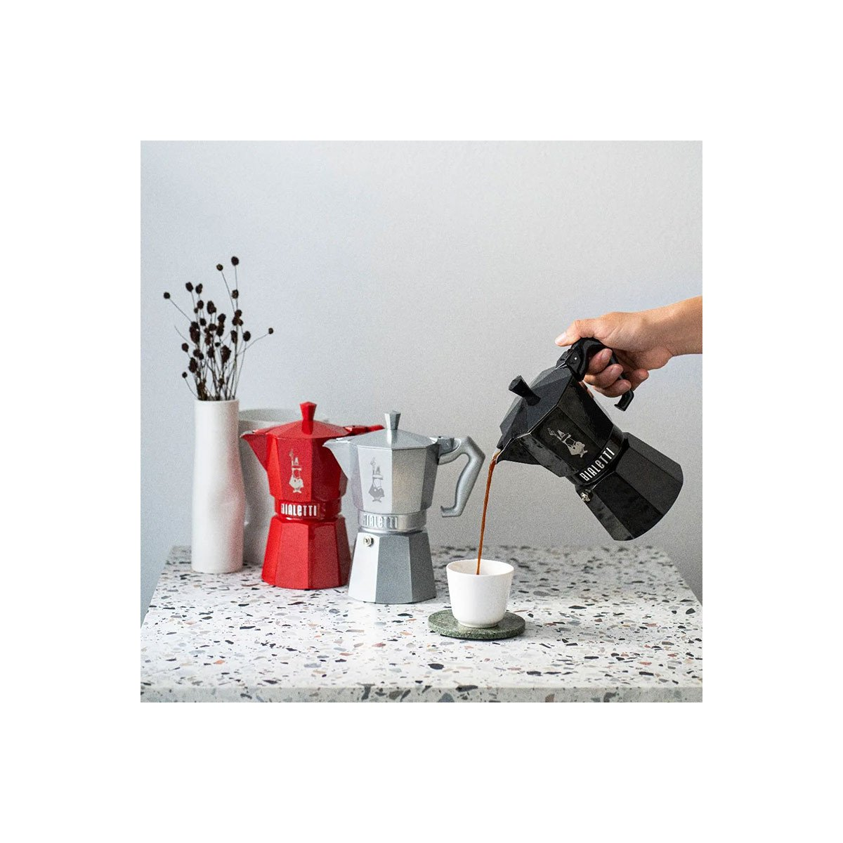 Una mano versa il caffè da una moka nera in una tazza, accanto ad altre moka su un tavolo.