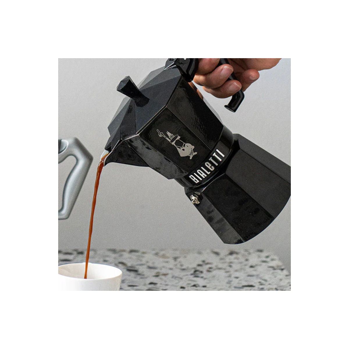 Una moka Bialetti nera versa il caffè in una tazza bianca.