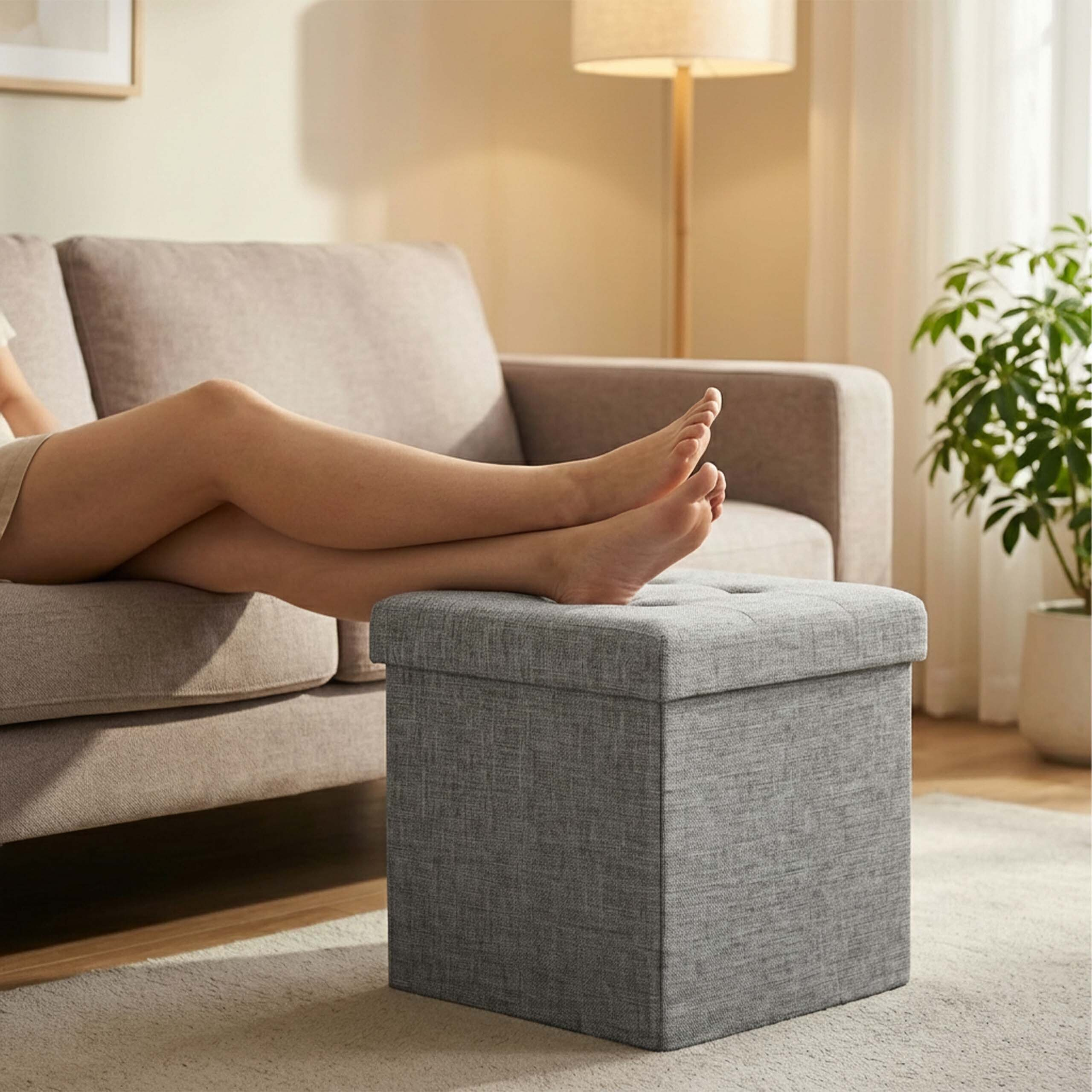 Füße ruhen auf einem grauen Hocker vor einem beigen Sofa im hellen Zimmer.