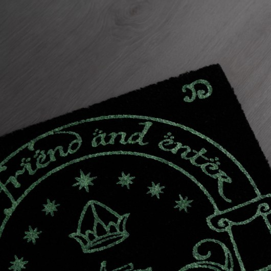 Un felpudo negro con estampado verde que incluye texto, estrellas y una corona, sobre suelo de madera gris.