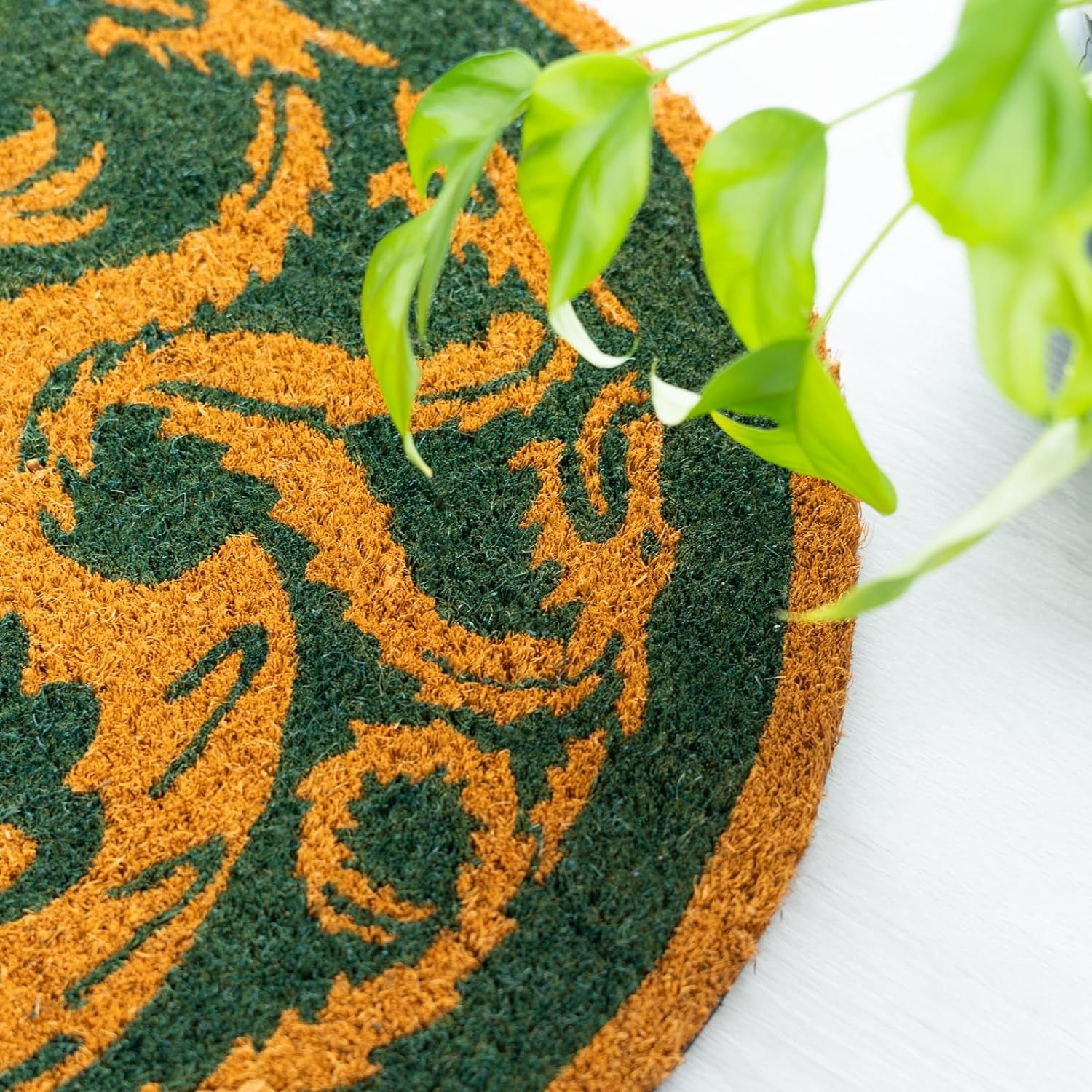 Uno zerbino rotondo verde e marrone con motivo a spirale, parzialmente coperto da foglie di piante verdi.