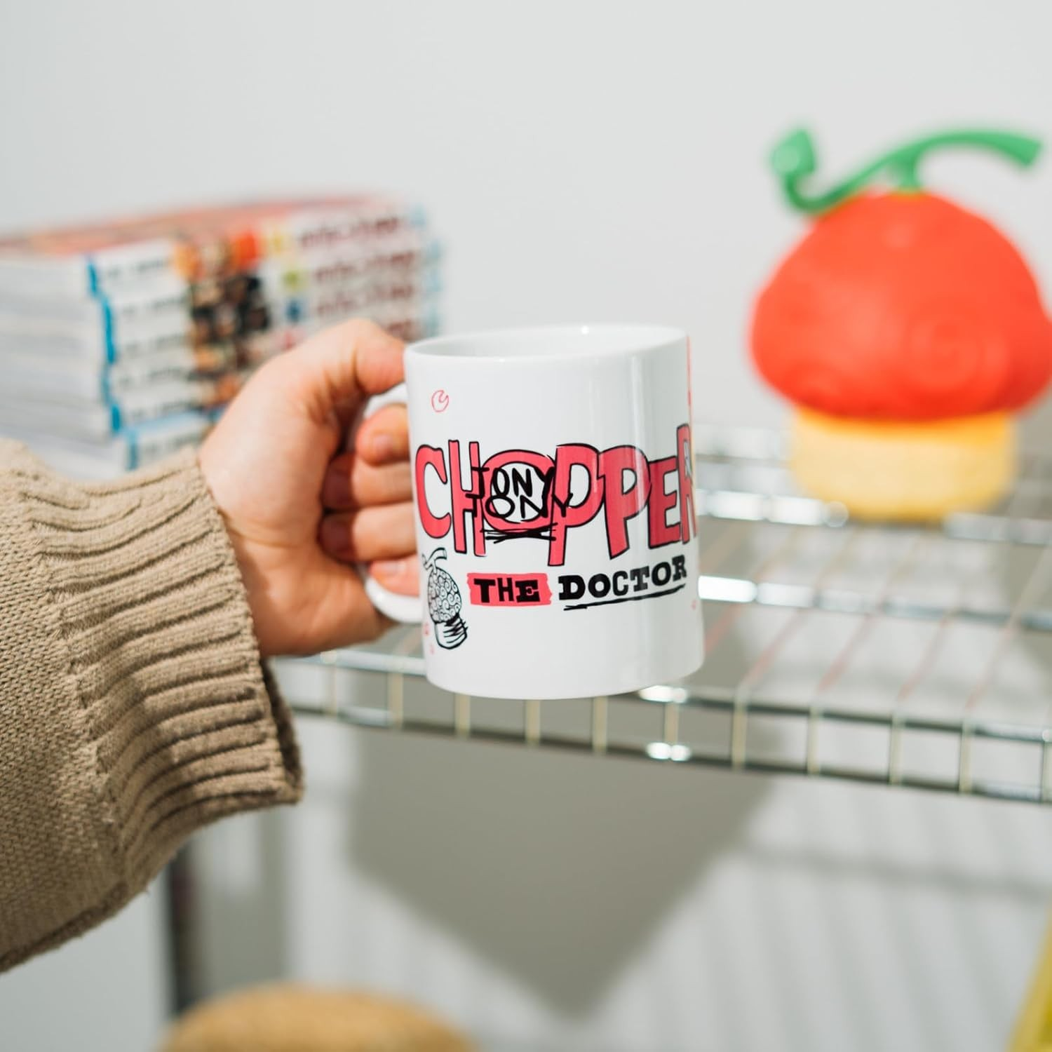 Una mano con un jersey beis sostiene una taza blanca con texto frente a una estantería.