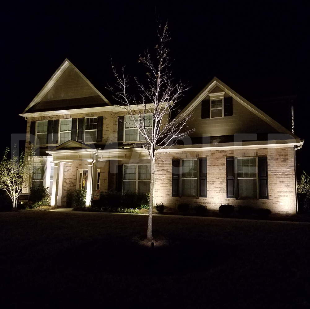 Una casa de ladrillo de dos pisos por la noche, iluminada con luces exteriores y un árbol sin hojas en el jardín.