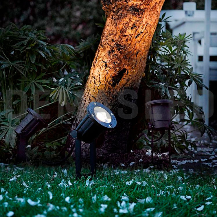 Tres focos de jardín negros iluminan el tronco de un árbol en un jardín por la noche.