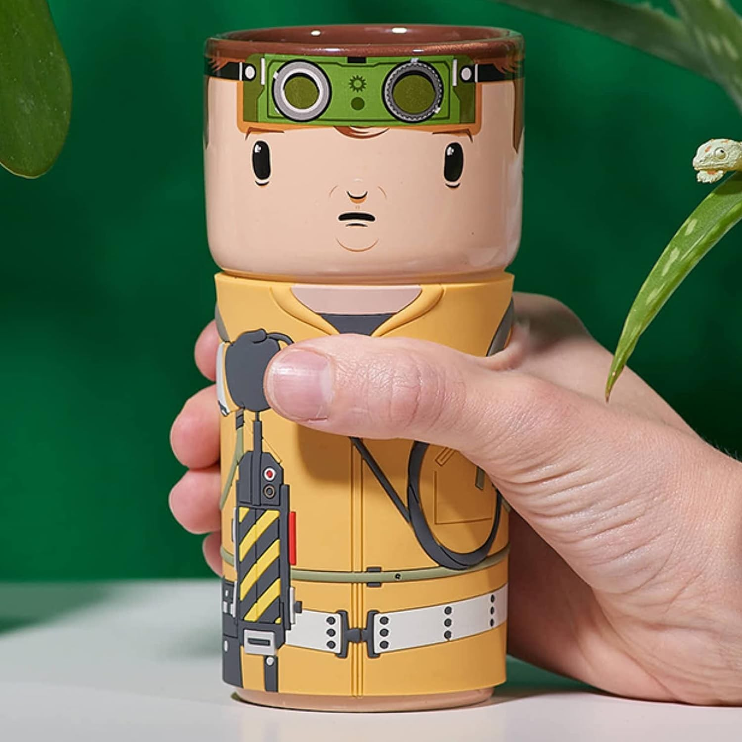 Eine Hand hält einen gelben Becher mit dem Bild eines Charakters mit Schutzbrille vor grünem Hintergrund.