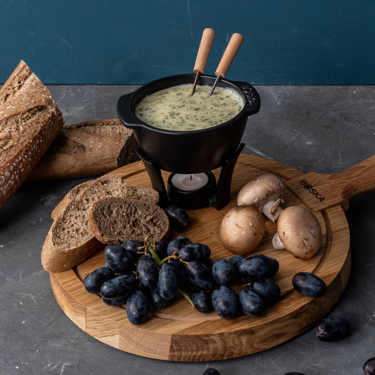 Käsefondue in einem schwarzen Topf auf einem Holzbrett, serviert mit Brot, Pilzen und Trauben.