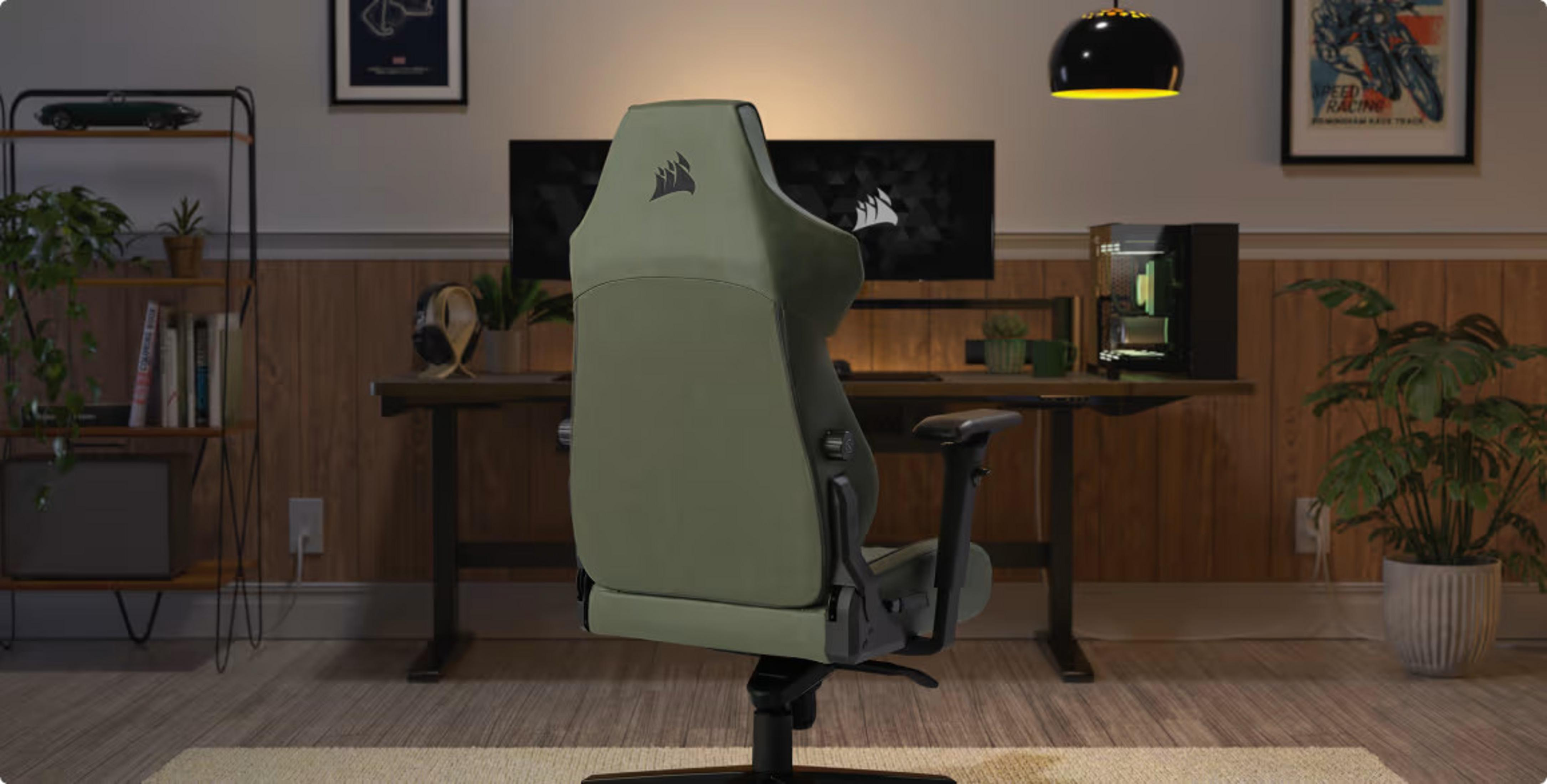 Ein olivgrüner Gaming-Stuhl steht im Vordergrund eines Raumes mit Schreibtisch, Monitor und Pflanzen.