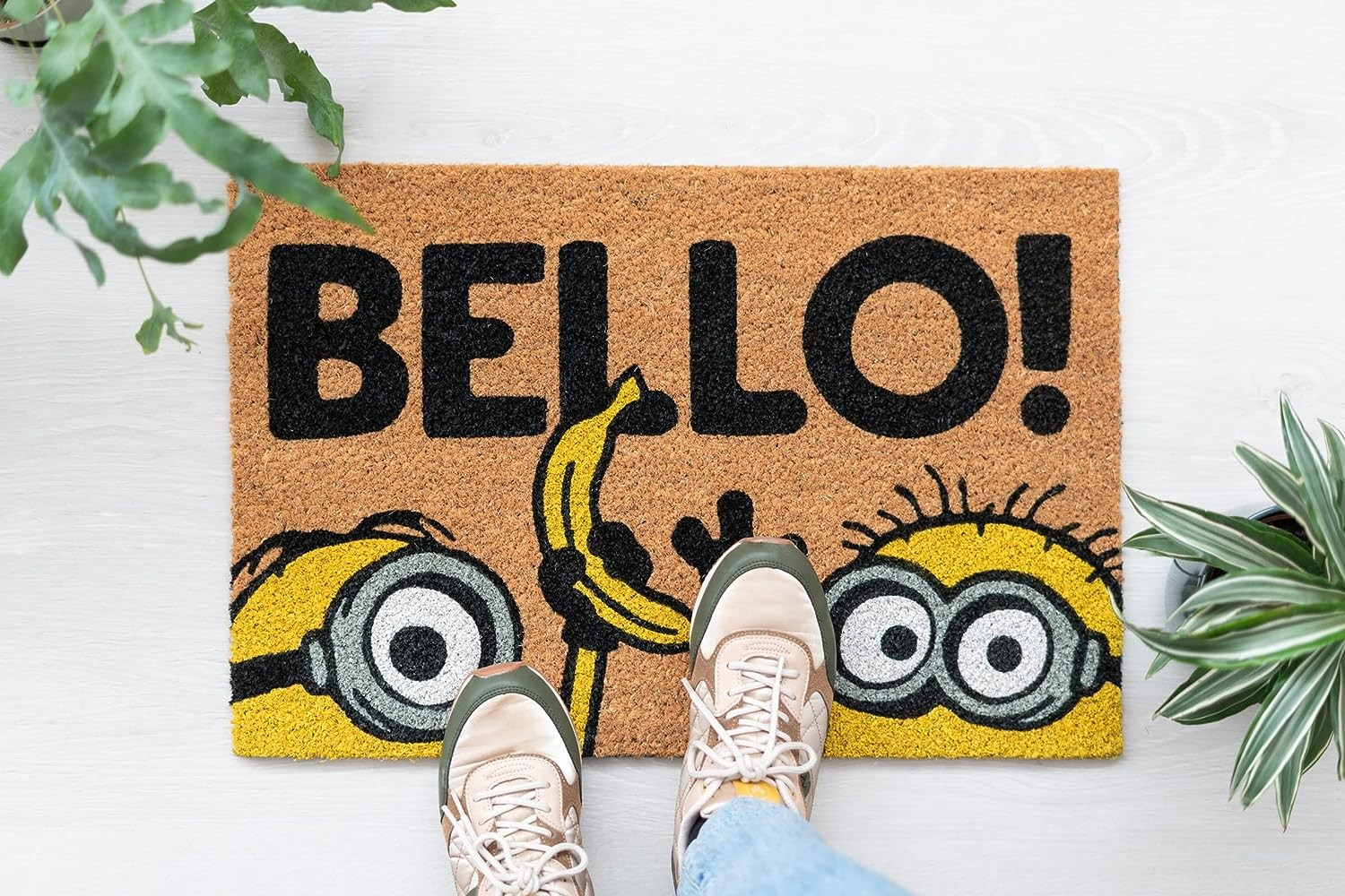 Un tappetino a tema Minions con la scritta BELLO! su pavimento in legno chiaro, affiancato da piante verdi.